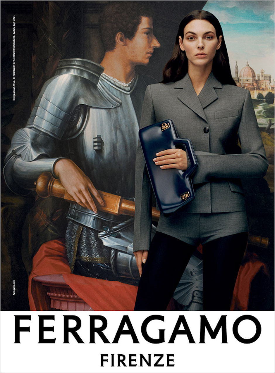 Modelka na tle obrazu Giorgio Vasariego z kampanii Ferragamo AW2023 ubrana w szary kostium z krótkimi spodenkami i dopasowaną szarą marynarką, w ręku trzyma niebieską torebkę kopertówkę. 